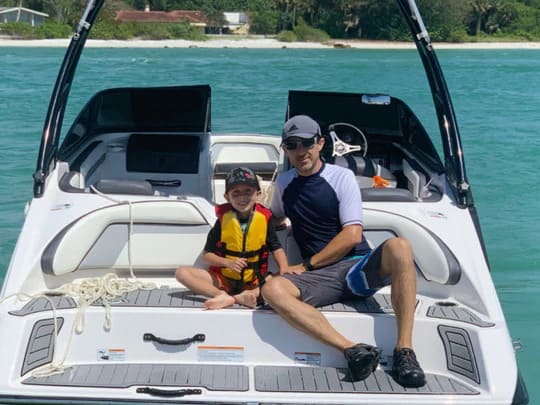 Man and child sitting on a boat in clear blue water with a beach in the background